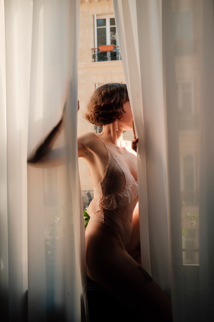Das Foto zeigt eine junge Frau, die in einem Fenster sitzt und die Vorhänge hält.